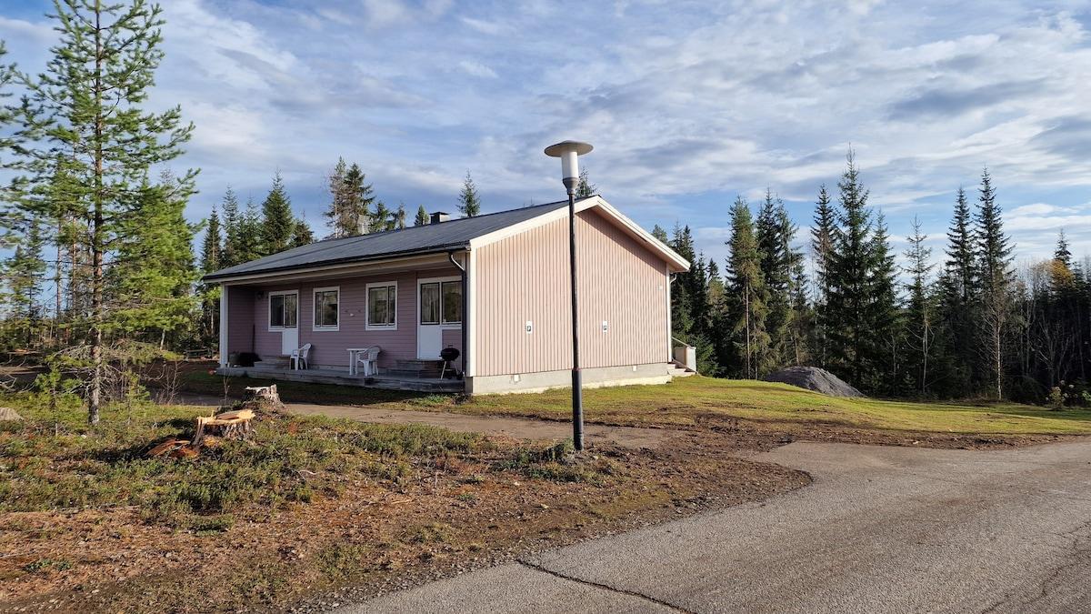 Ljust trähus med sadeltak och veranda i en skogsmiljö.